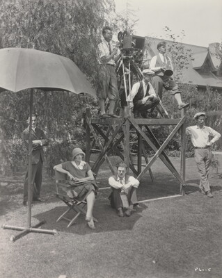 A black-and-white photograph of three people and a movie camera on a camera riser, two people standing next to the riser, and two people seated beneath a large umbrella.