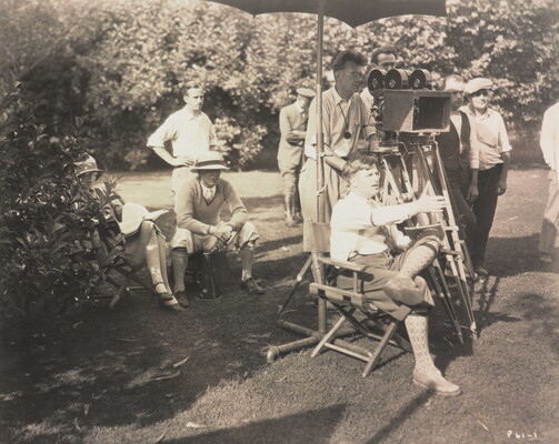 A black-and-white photograph of a group of White men, including a seated director gesturing in front of him, a camera man, and others in the background.