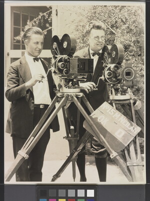 A black-and-white photograph of two White men standing next to each other behind two large video cameras.