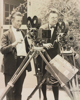 A black-and-white photograph of two White men standing next to each other behind two large video cameras.