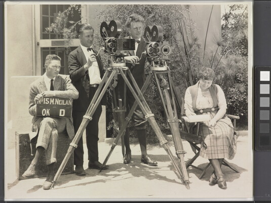 A black-and-white photograph of three White men, two standing behind video cameras, one seated with a clapperboard, and a White woman seated in a folding chair.