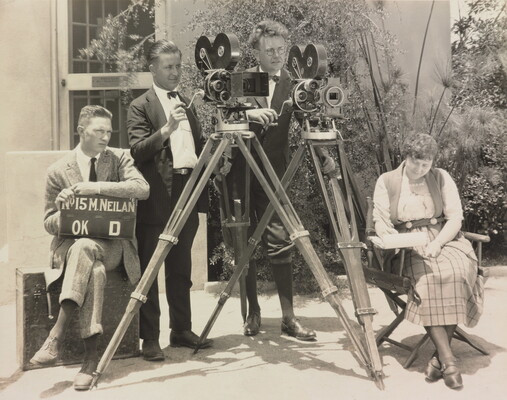 A black-and-white photograph of three White men, two standing behind video cameras, one seated with a clapperboard, and a White woman seated in a folding chair.