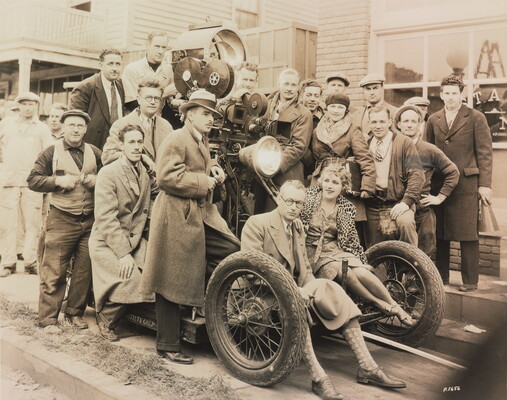 A sepia-toned photograph of a group of people gathered around a cart on which is propped a large movie camera.