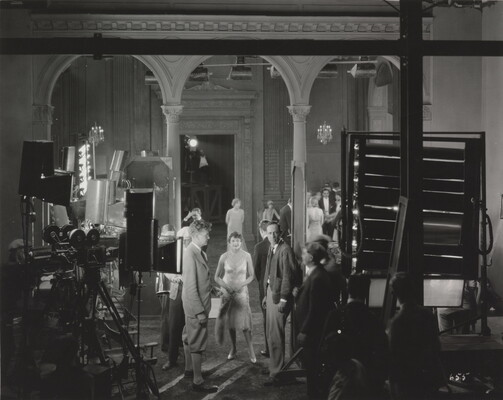 A black-and-white photograph of a group of well-dressed people standing around a movie set surrounded by film equipment.