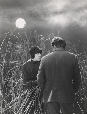 A black-and-white photograph of the back of a man facing a White woman wearing a coat and hat in a field of high grass under a full moon.