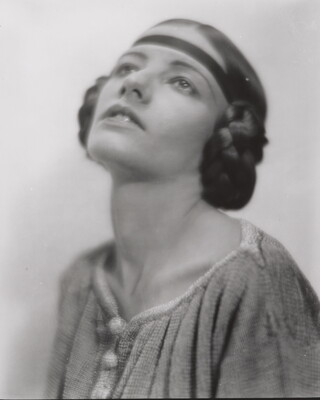 A black-and-white portrait of a White woman, head angled up, with her hair braided into buns on the sides of her head.