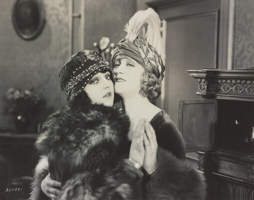 A black-and-white photograph of two White women, both wearing hats and fur coats, embracing each other with their cheeks touching and holding hands.