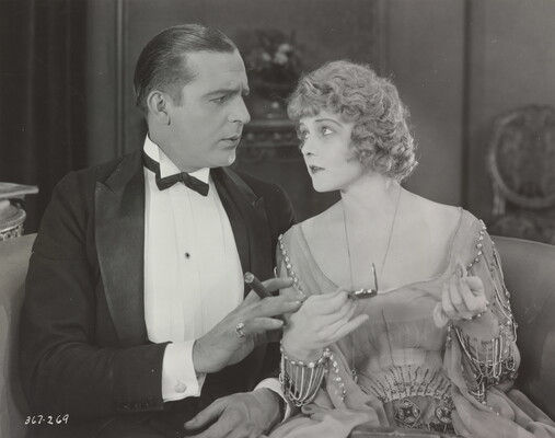 A black-and-white photograph of a White man in a suit holding a cigar sitting next to a White woman in a beaded formal dress, both looking at each other.