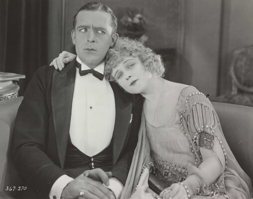 A black-and-white photograph of a White man in a suit sitting next to a White woman in a formal beaded dress who is leaning her head on the man's shoulder.