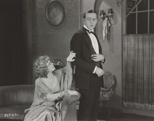 A black-and-white photograph of a seated White woman wearing a formal beaded dress who is clutching the arm of a standing White man wearing a suit.