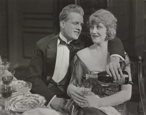 A black-and-white photograph of a seated White man with his arm around the shoulder of a White woman in a formal dress; he is pouring her a drink with the arm he has around her.