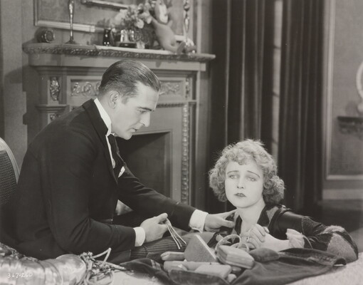A black-and-white photograph of a White man, one hand on the shoulder of a White woman, as he offers her jewelry with the other hand.