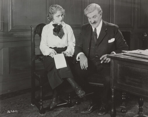 A black-and-white photograph of a young White woman wearing a blouse and skirt and an older White man in a three-piece suit seated next to each other; the woman holds a notebook on her lap.