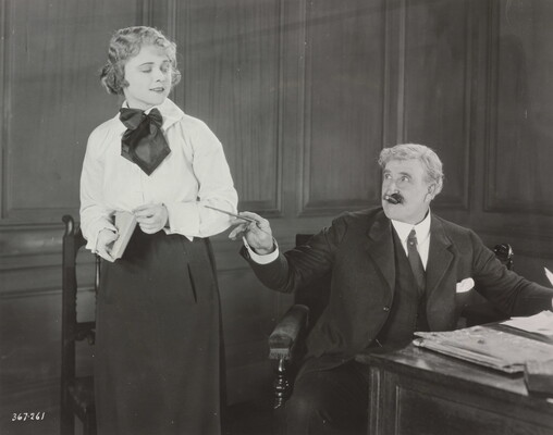 A black-and-white photograph of young White woman wearing a blouse and skirt and holding a notebook standing next to an older White man in a three-piece suit seated at a desk.
