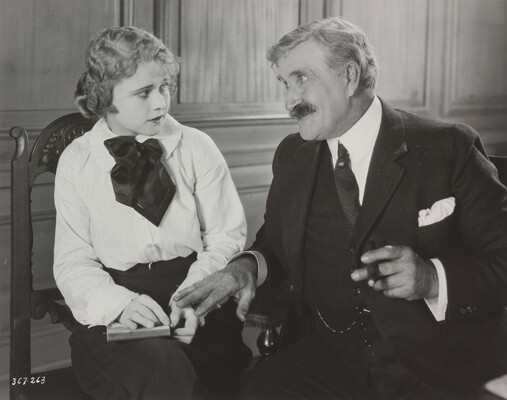 A black-and-white photograph of a young White woman wearing a blouse and skirt holding a notebook on her lap and an older White man in a three-piece suit leaning toward the woman.