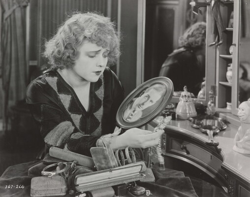 A black-and-white photograph of a White woman, seated at a vanity, gazing down at a photograph of a White man in an oval frame.