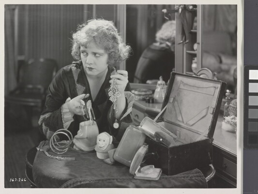 A black-and-white photograph of a White woman seated at a vanity, holding two handfuls of jewelry and looking off to the side.