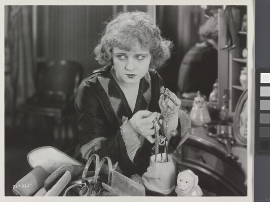 A black-and-white photograph of a White woman seated at a vanity, holding two handfuls of jewelry and looking off to the side.