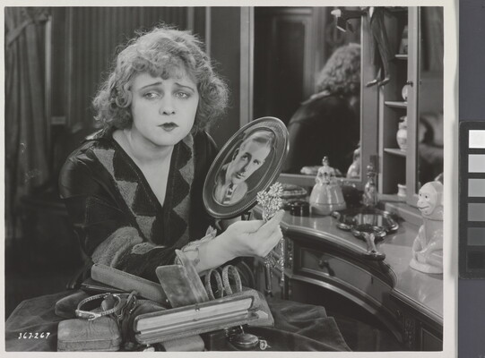 A black-and-white photograph of a White woman sitting at a vanity holding a brooch and a photograph of a White man as she gazes into the distance.