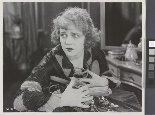 A black-and-white photograph of a White woman seated at a vanity clutching a variety of objects and jewelry to her chest.