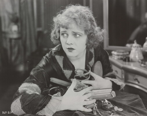 A black-and-white photograph of a White woman seated at a vanity clutching a variety of objects and jewelry to her chest.