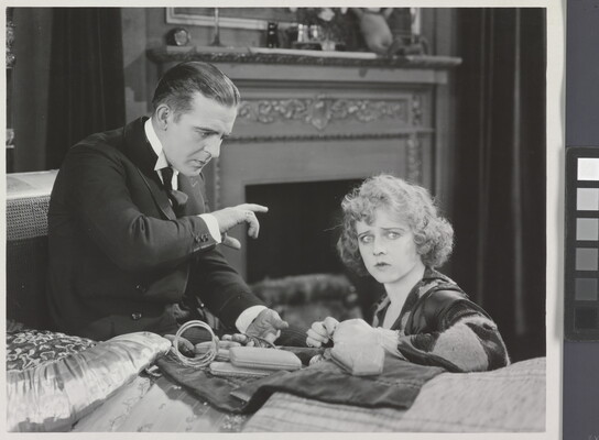 A black-and-white photograph of a White man gesturing toward a seated White woman who is looking toward the viewer.