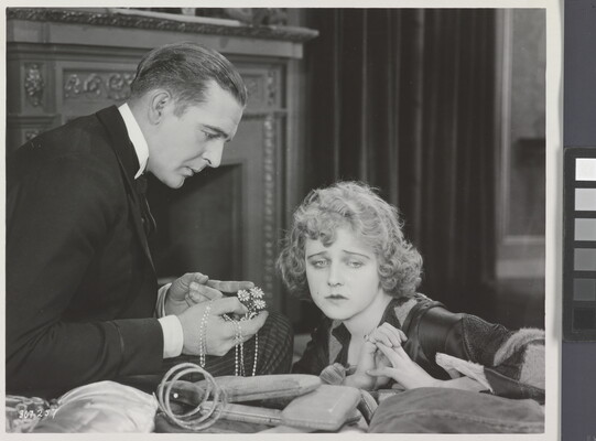 A black-and-white photograph of a White man presenting a White woman with jewelry as she sadly gazes into the distance.