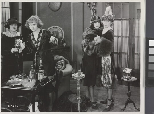 A black-and-white photograph of four White women standing in a room looking at something in the distance, two of the women holding each other in surprise.
