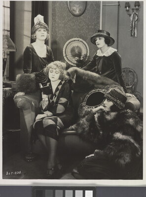 A black-and-white photograph of four elaborately-dressed White women, two standing, one reading on a sofa, the other seated on the floor.