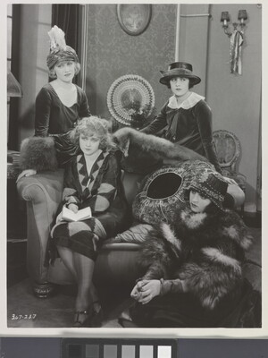 A black-and-white photograph of four elaborately-dressed White women, two standing, one on a sofa with a book in her lap, the other seated on the floor.