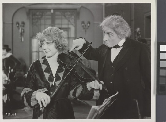 A black-and-white photograph of a White woman playing a violin and an older White man next to her with his finger violin's chinrest.