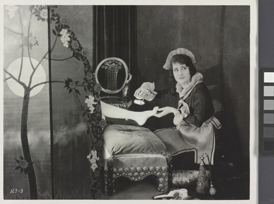 A black-and-white photograph of a White maid giving a pedicure to the foot of a woman who is hidden behind a screen.