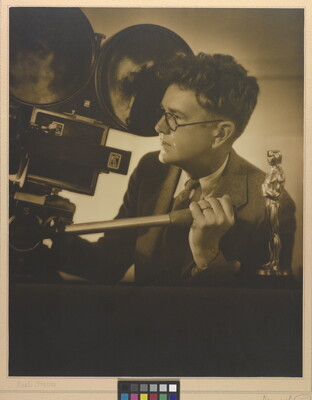 A sepia-toned photograph of a White man looking into a large movie camera and an Oscar statuette on a table next to him.
