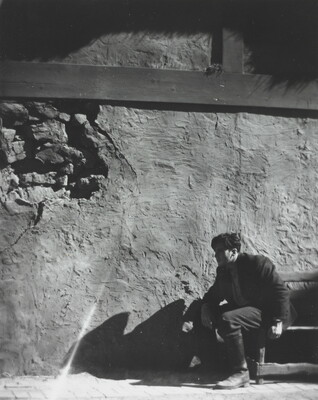 A black-and-white photograph of a man wearing a hat crouched next to a large rock wall.