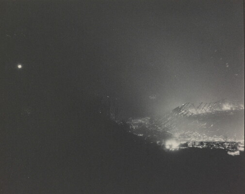 A black-and-white photograph of a large wooden structure, partially hidden by a hill, burning at night.