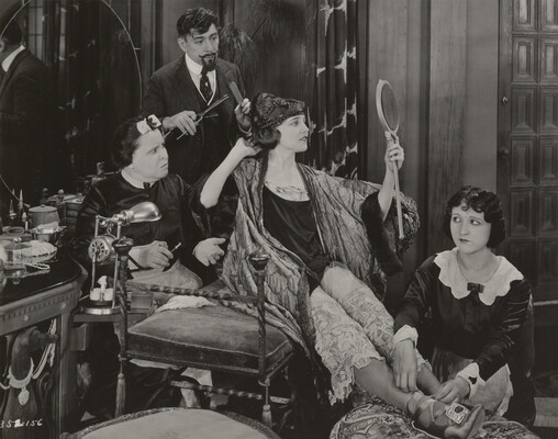 A black-and-white photograph of an elaborately-dressed White woman seated on a bench, gazing into a hand mirror, surrounded by people who are helping her dress.