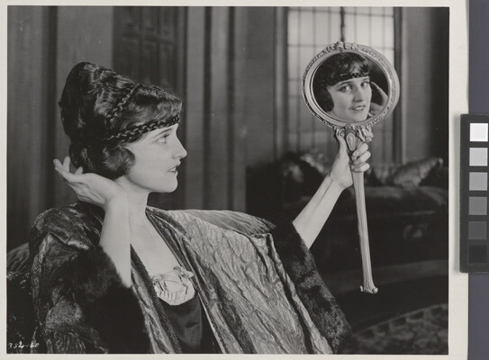 A black-and-white photograph of an elaborately dressed White woman holding up a mirror and gazing into it, her gaze in the mirror meeting the viewer's eye.