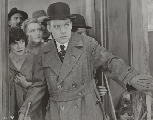 A black-and-white photograph of a White man wearing a bowler hat and a long, double-breasted overcoat opening a door; he and the group of people behind him look surprised.