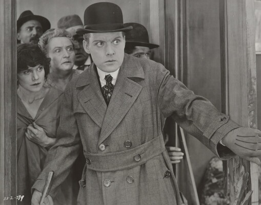 A black-and-white photograph of a White man wearing a bowler hat and a long, double-breasted overcoat opening a door; he and the group of people behind him look surprised.
