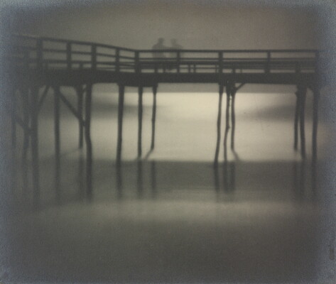 A black-and-white soft-focus photograph of a wooden dock over water; the shadows of two people stand on the dock.