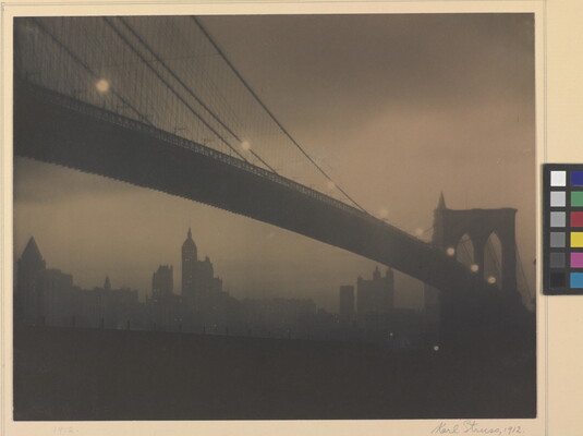 A sepia-toned photograph of a long bridge with a foggy cityscape in the background taken at night.