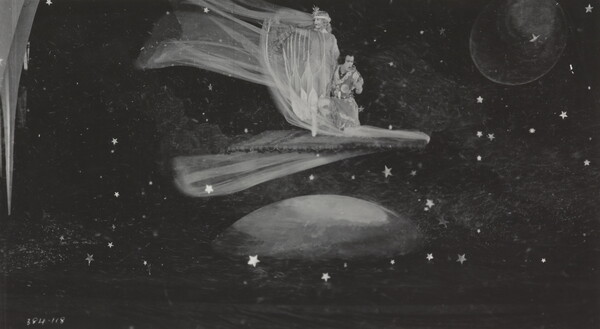 A black-and-white photograph of a White woman wearing a flowing dress and a White man kneeling next to her on a flying carpet traveling through space.