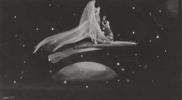 A black-and-white photograph of a White woman wearing a flowing dress and a White man kneeling next to her on a flying carpet traveling through space.