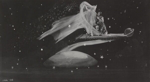 A black-and-white photograph of a White woman wearing a flowing dress and a White man kneeling next to her on a flying carpet traveling through space.