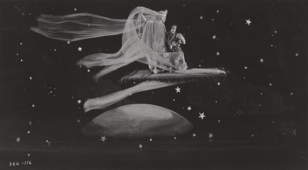 A black-and-white photograph of a White woman wearing a flowing dress and a White man kneeling next to her on a flying carpet traveling through space.