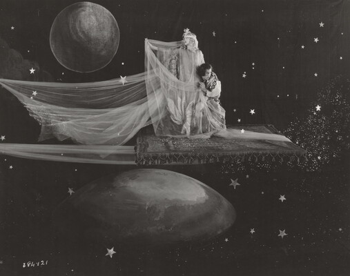 A black-and-white photograph of a White woman wearing a flowing dress and a kneeling White man holding on to her on a flying carpet traveling through space.