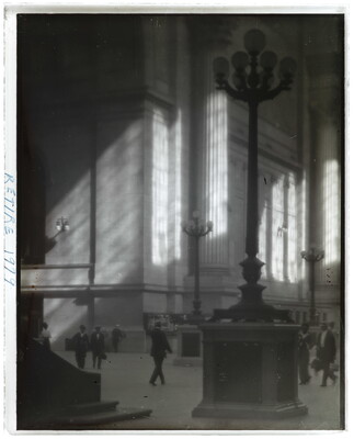 A black-and-white photograph of people walking through a sunlit plaza with towering light posts and columns.