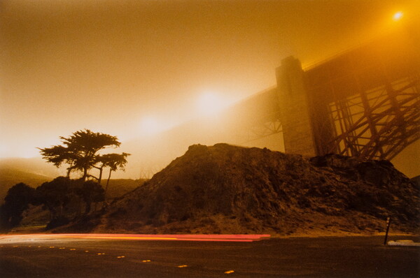 A color photograph of the underside of a bridge from a road next to hills and trees; sunlight and lights on the bridge give the image an orange glow.