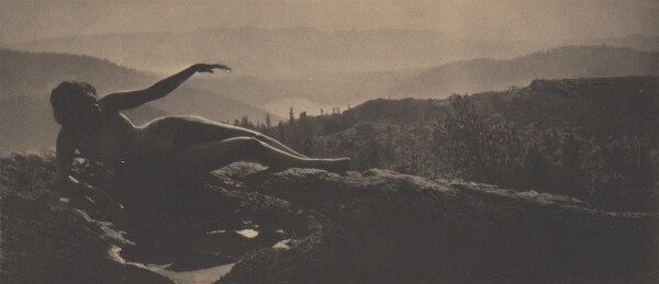 A sepia-toned photograph of a nude woman in shadow lying on a hilltop with cascading hills behind her.