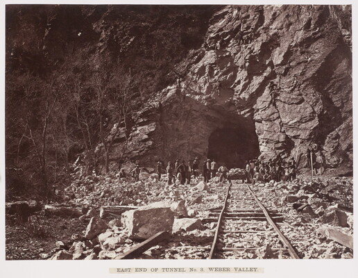 A black-and-white photograph of railroad tracks leading into a rocky tunnel, with workers gathered at the  tunnel entry.